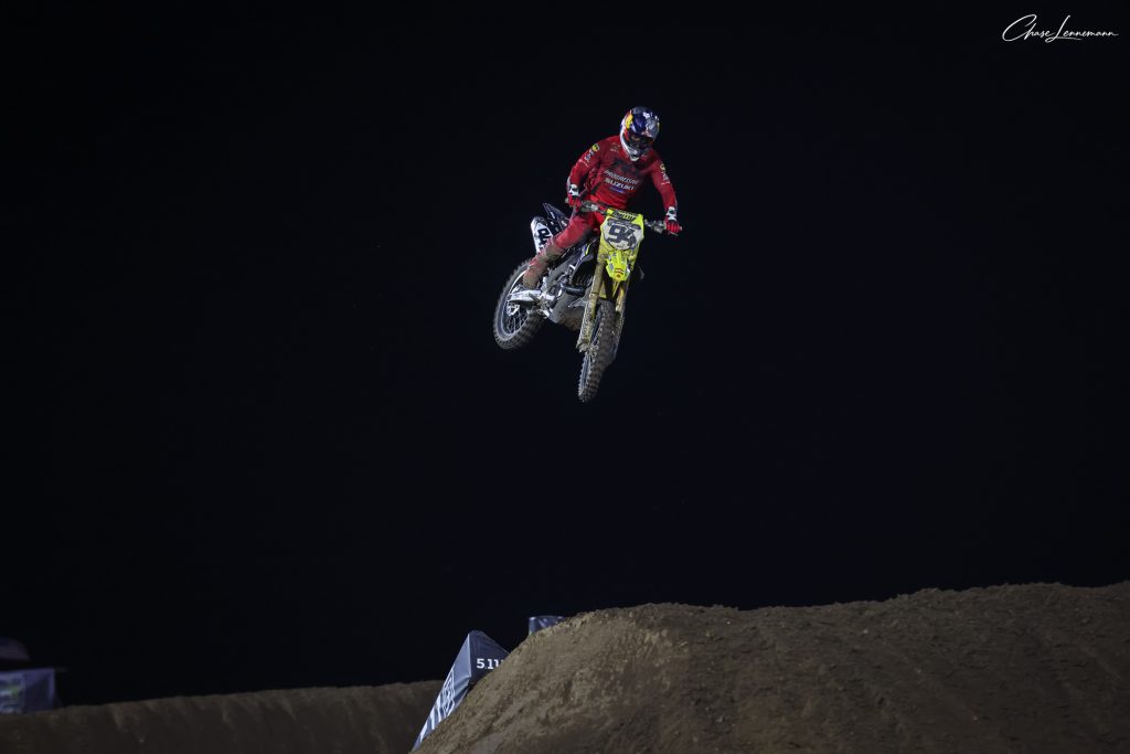 Kyle Chisholm jumps a yellow motocross bike above a dirt mound against a dark background.