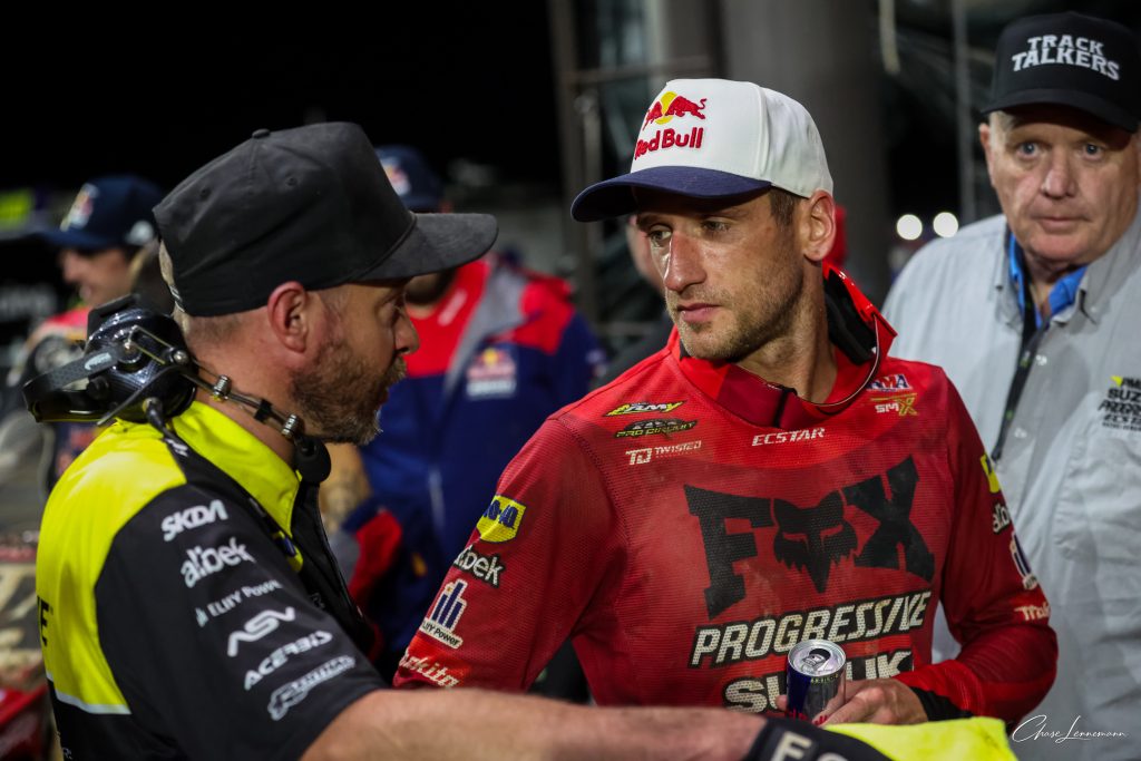 A man in a red racing jersey and Red Bull cap talks with a crew member wearing a headset around his neck.
