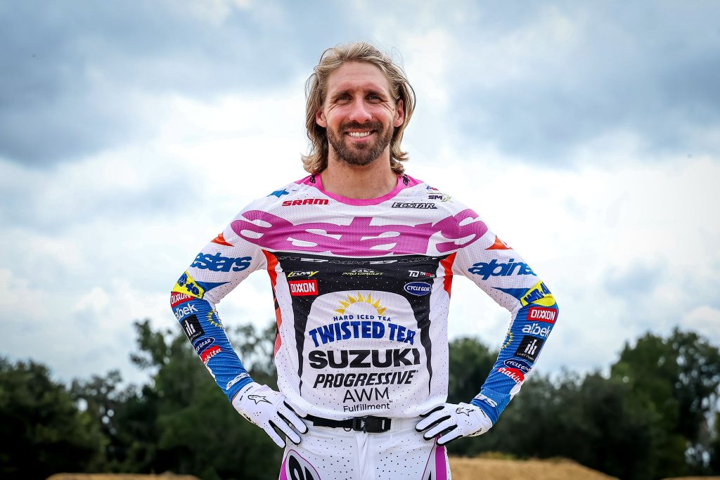 A smiling motocross rider in pink, white and blue racing gear stands with hands on hips beneath a cloudy sky.